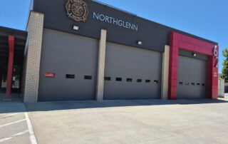 Northglenn Fire Station Exterior Dark gray painted commercial fire station facade with cream brick pillars, red metal accent panels, and large sectional bay