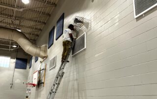 Gymnasium Interior Wall Painting Painter on tall ladder applying white paint to brick gymnasium wall with blue sound-dampening panels and industrial ceiling