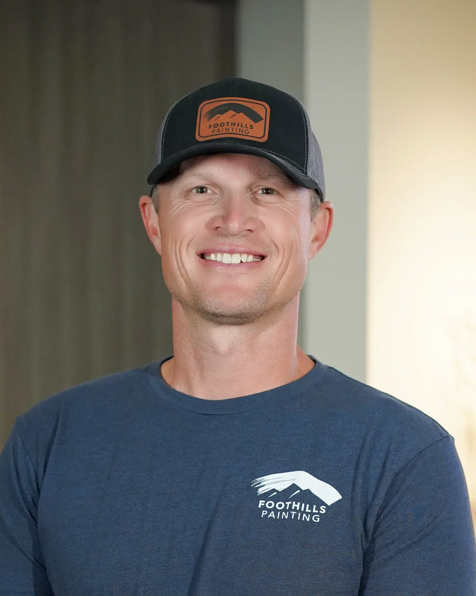 Foothills Painting Team Member Smiling man wearing navy Foothills Painting branded t-shirt and black mesh cap with orange logo against neutral wall