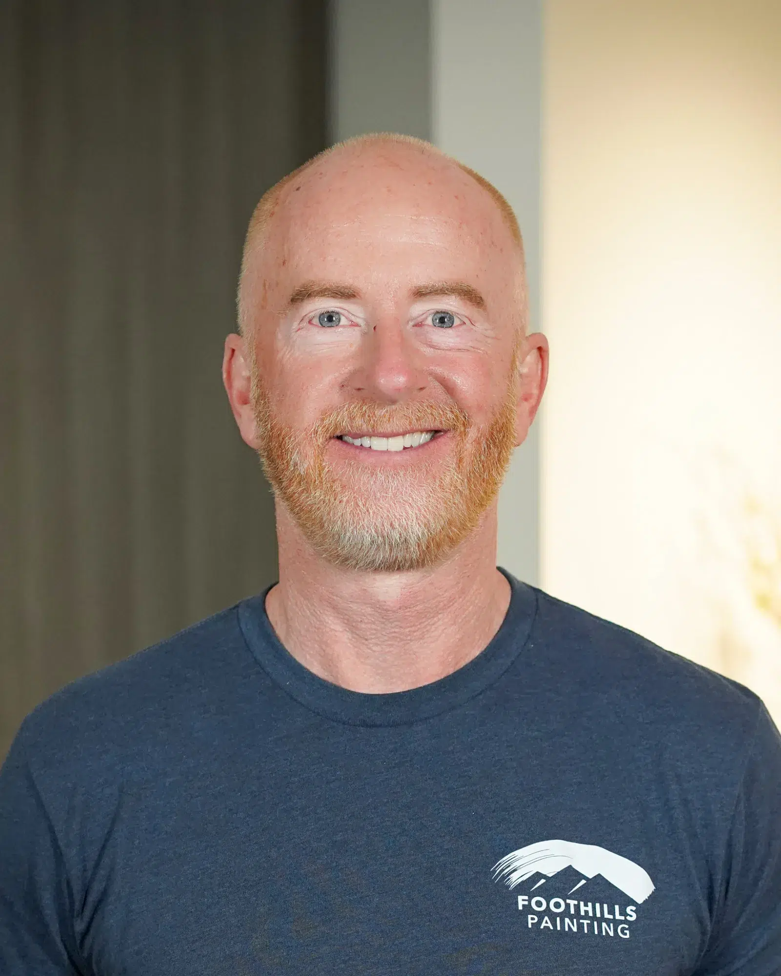 Foothills Painting Team Member Smiling man with reddish-blonde beard wearing dark navy Foothills Painting branded t-shirt against neutral wall