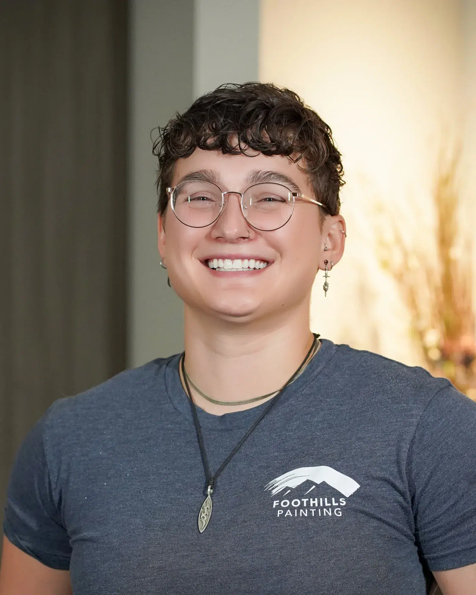 Foothills Painting Team Member Smiling person with short dark curly hair, rose-tinted glasses, and silver earrings wearing dark gray Foothills Painting