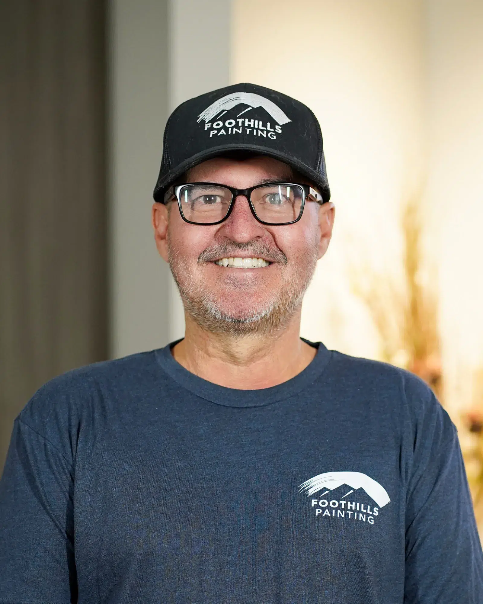 Foothills Painting Team Member Smiling man with gray beard wearing dark navy Foothills Painting branded cap and t-shirt with glasses against neutral wall