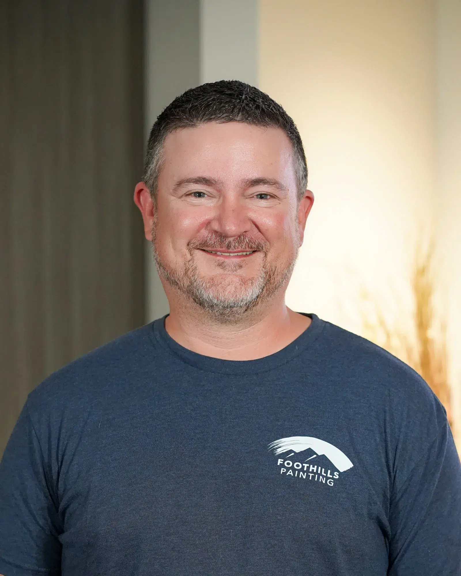 Foothills Painting Team Member Smiling man with brown beard wearing dark navy Foothills Painting branded t-shirt against neutral wall backdrop