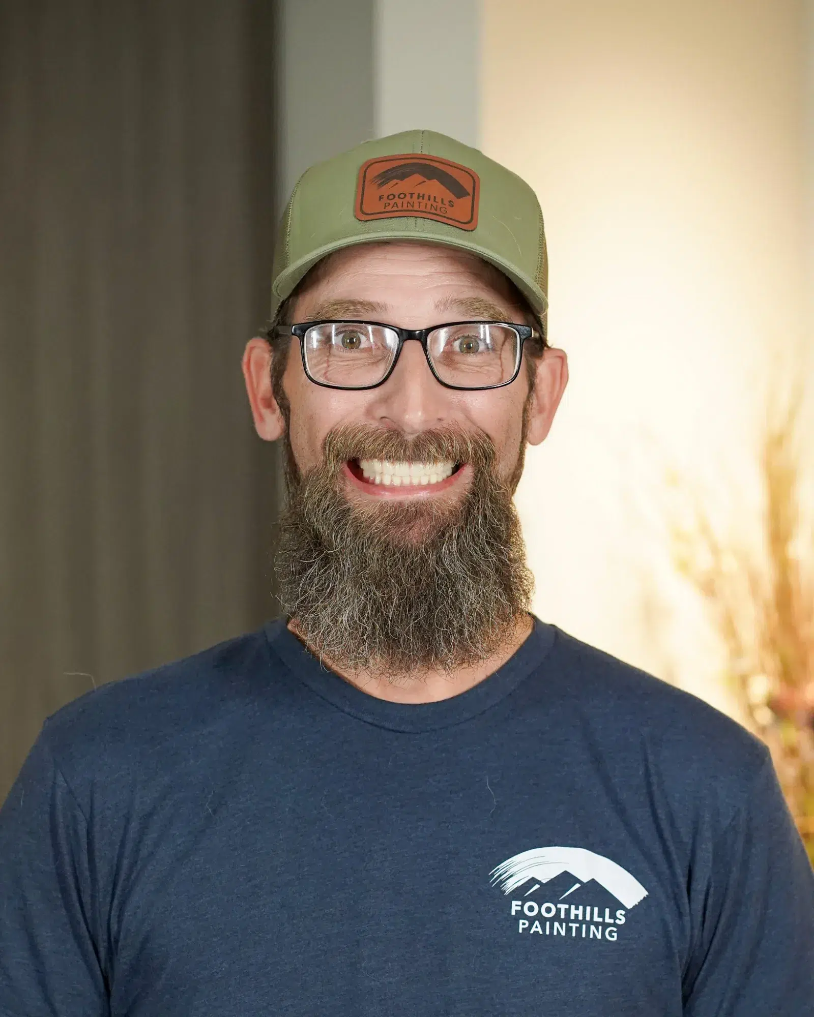 Foothills Painting Team Member Smiling man with full dark beard wearing sage green Foothills Painting cap, black-framed glasses, and navy branded t-shirt