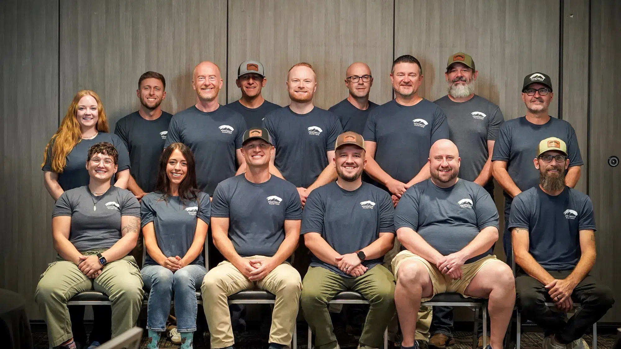 Foothills Painting Team Portrait Fourteen smiling team members wearing dark gray branded shirts pose in two rows against textured gray wall backdrop
