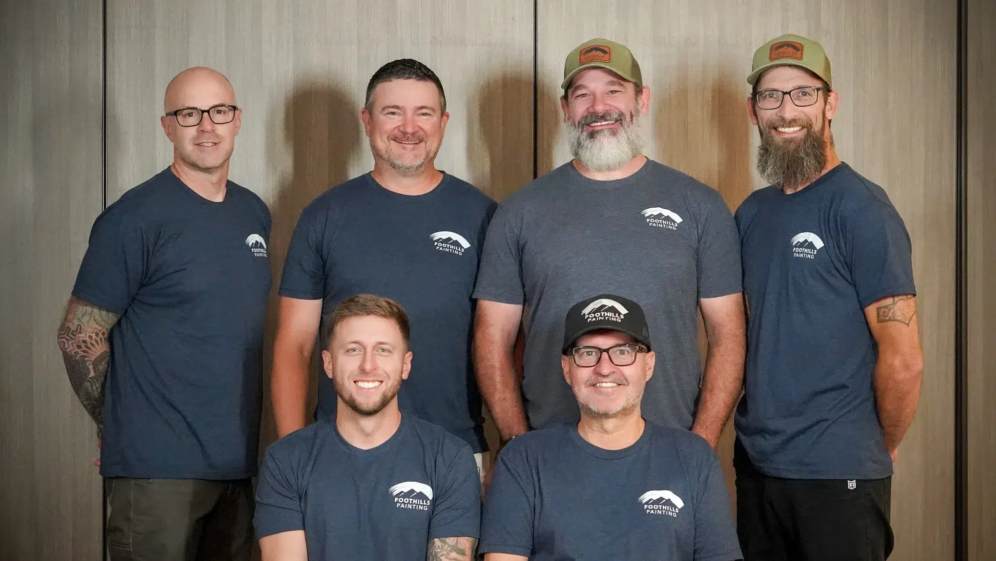 Foothills Painting Team Portrait Six smiling Foothills Painting team members wearing navy branded shirts pose against textured gray and tan wall backdrop