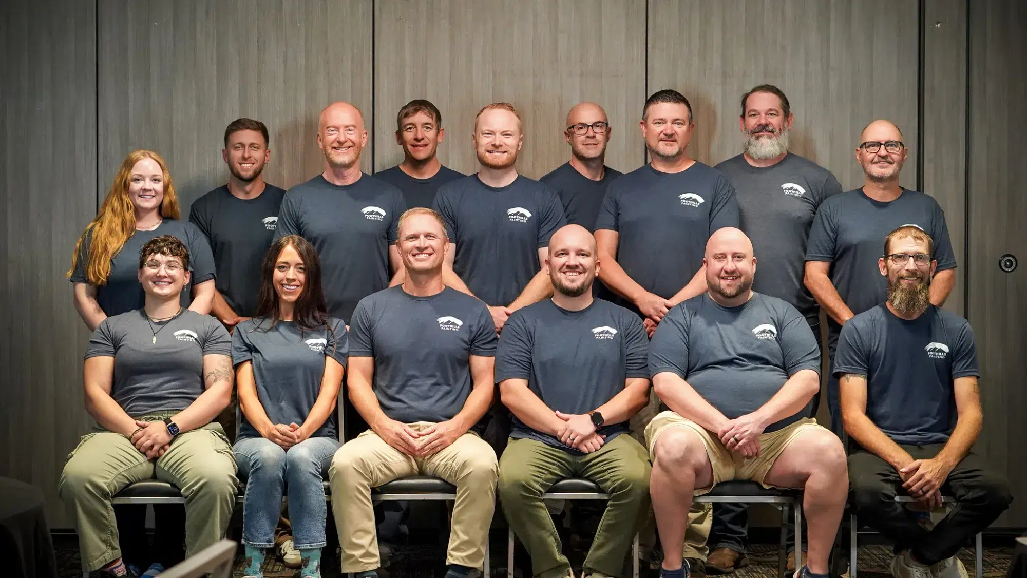 Foothills Painting Team Portrait Sixteen smiling team members wearing dark gray branded shirts pose in two rows against textured gray wall backdrop