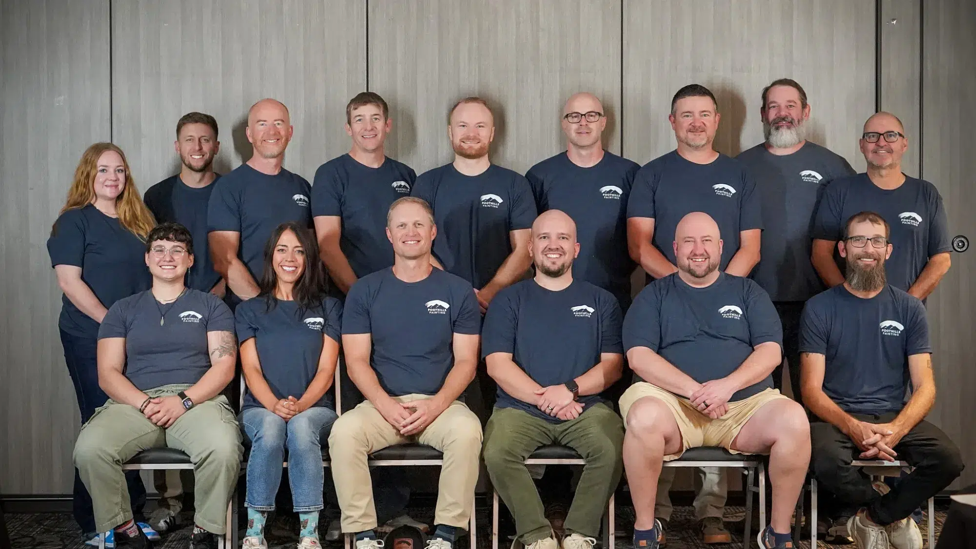 Foothills Painting Team Portrait Eighteen smiling team members wearing dark navy branded shirts pose in two rows against textured gray wall backdrop