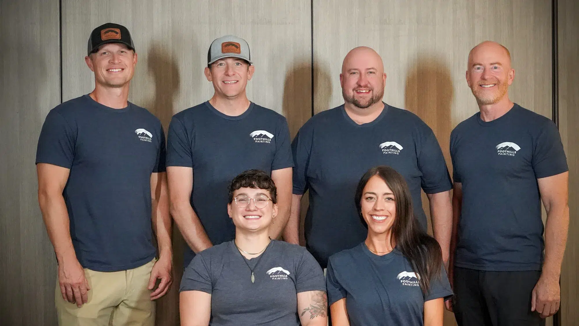Foothills Painting Team Portrait Six smiling team members wearing navy Foothills Painting branded shirts pose against neutral textured wall backdrop