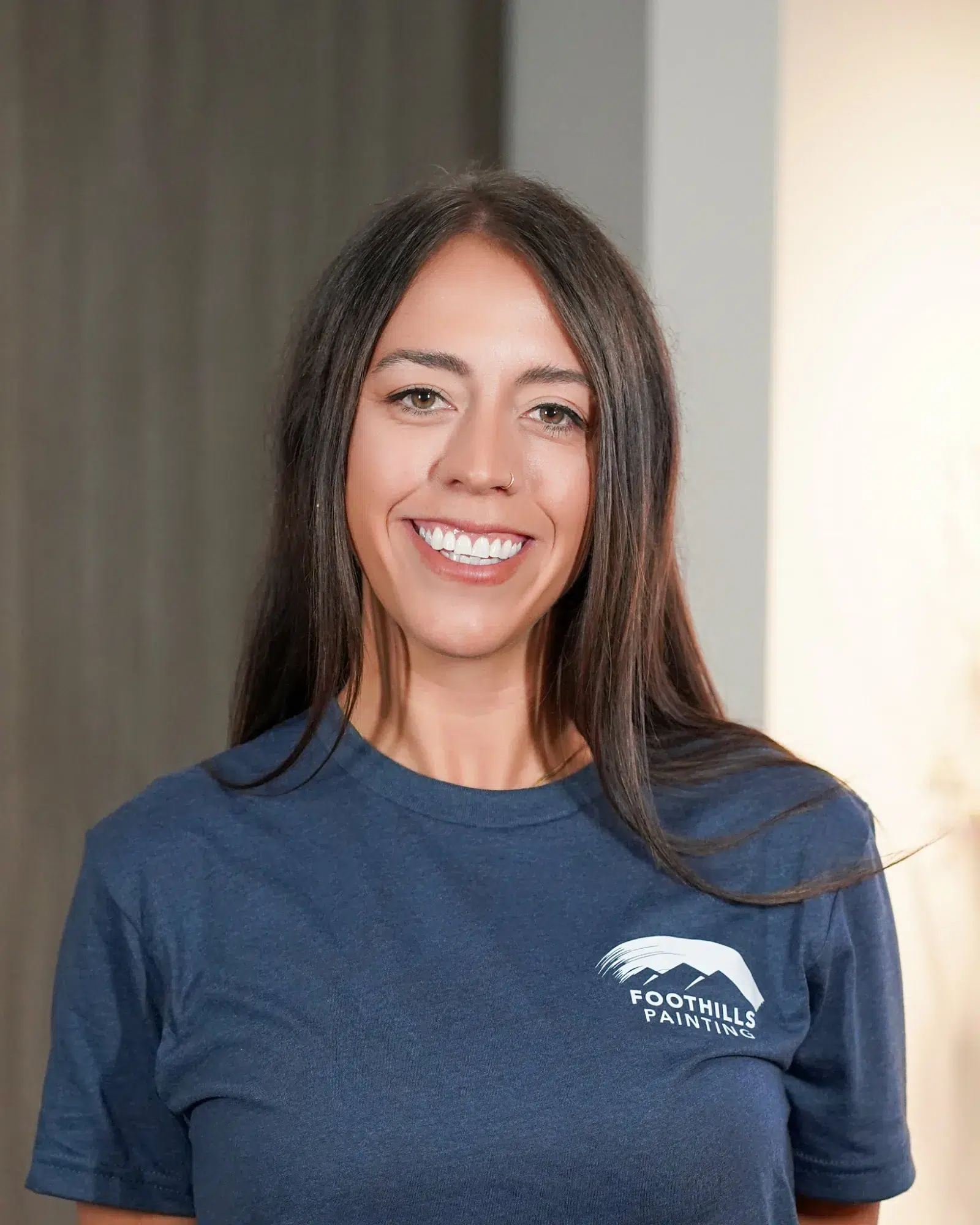 Foothills Paintings Team Member Smiling woman with long dark hair wearing navy Foothills Paintings branded t-shirt against neutral striped wall